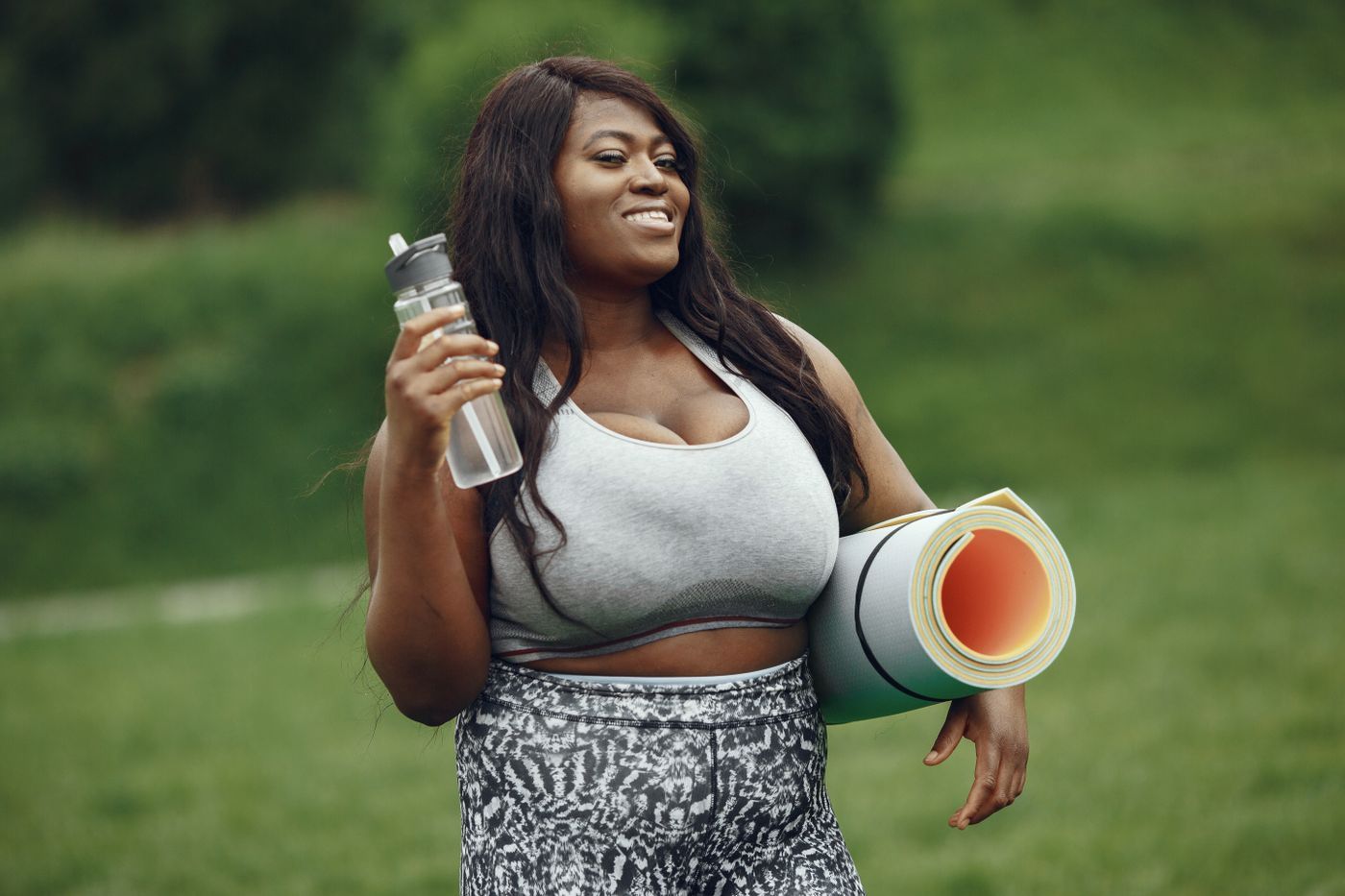 A smiling woman outdoors carrying an exercise mat and water bottle
