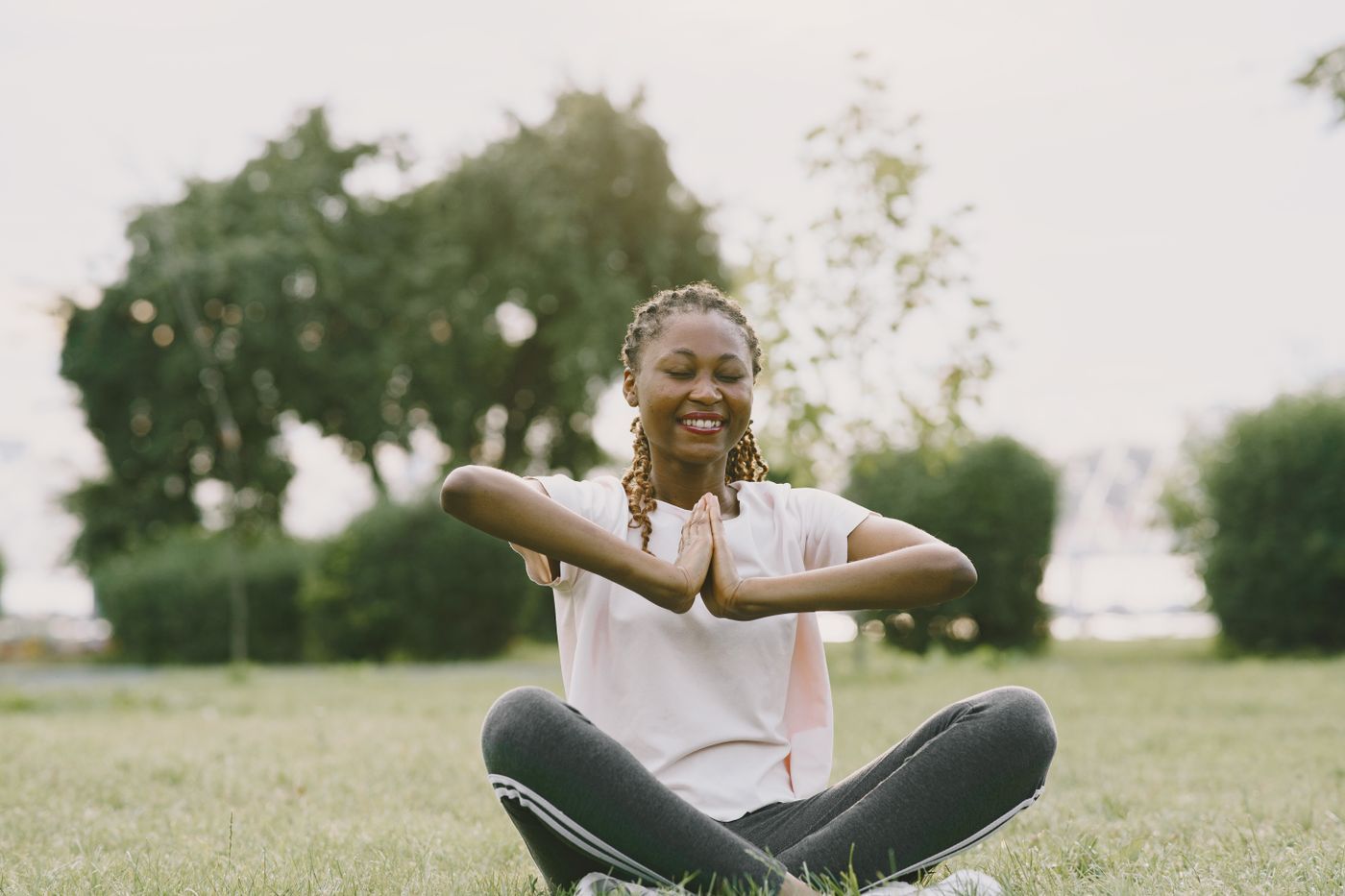 A woman sitting on the grass in a park, smiling with her eyes closed